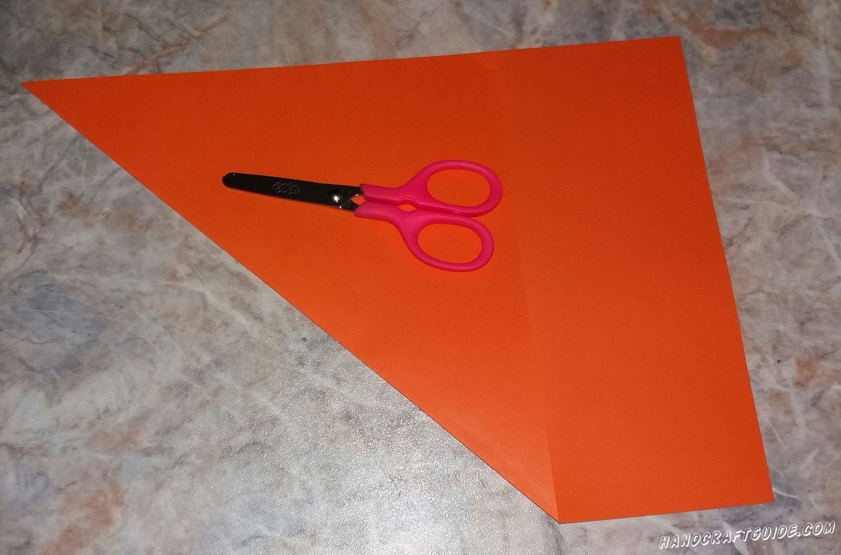 Начинаем мы с того что вырезаем ровный квадрат. Для этого берем оранжевый или красный лист бумаги и загибаем один угол. Всё что остается за треугольником мы обрезаем.