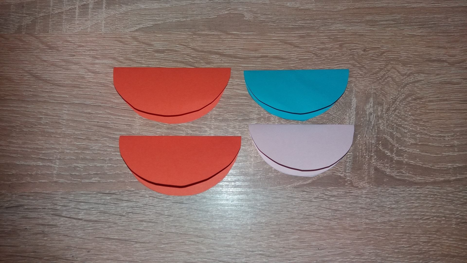 Для начала мы вырезаем 2 одинаковых красных круга, 1 розовый и 1 голубого цвета. Сгибаем эти кружки пополам