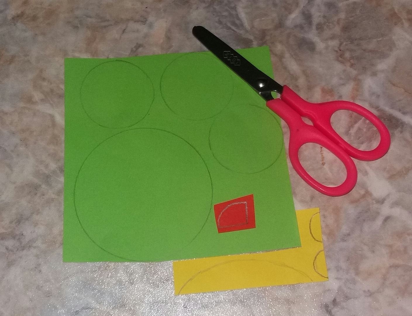 Для начала берем зеленую бумагу и вырезаем 1 большой и 3 меньших круга. Затем вырезаем длинный полуовал и 2 полукруга желтого цвета. Ну и в конце подготовки вырезаем красную детальку, как показано на фото.