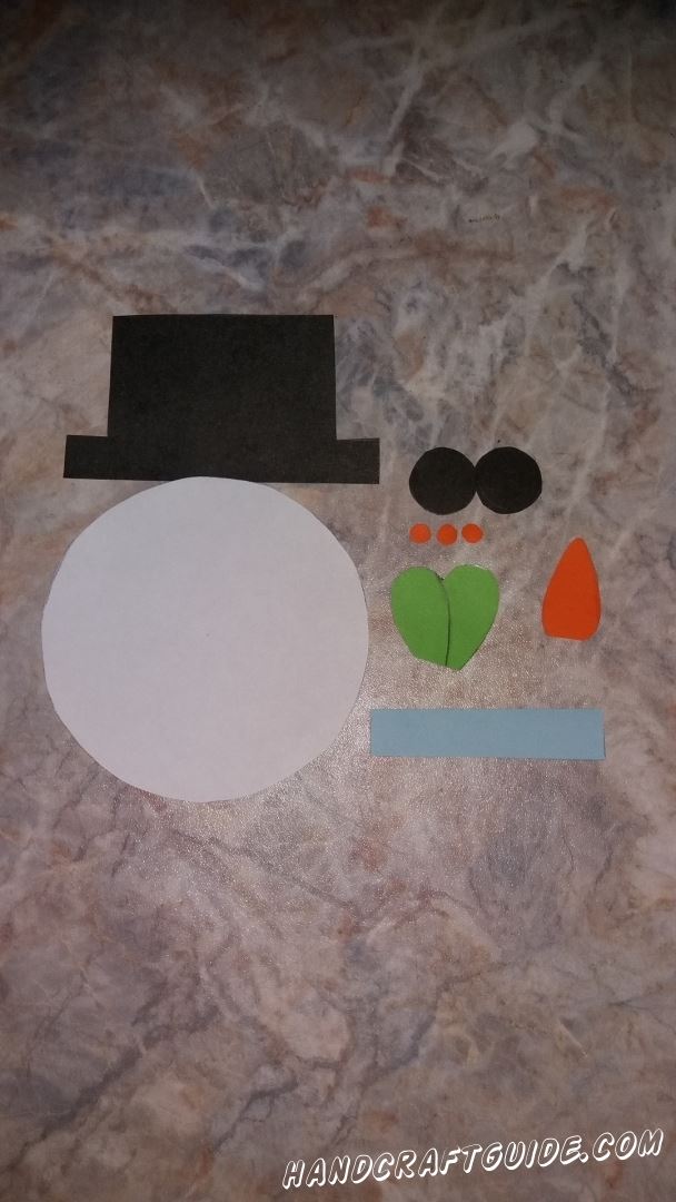 Сначала вырезаем все необходимые нам детали из цветной бумаги: большой белый круг, черная шляпа и 2 кружочка, голубая полоска, сердечко зелёного цвета, небольшая морковь и 3 очень маленьких кружочка оранжевого цвета. 