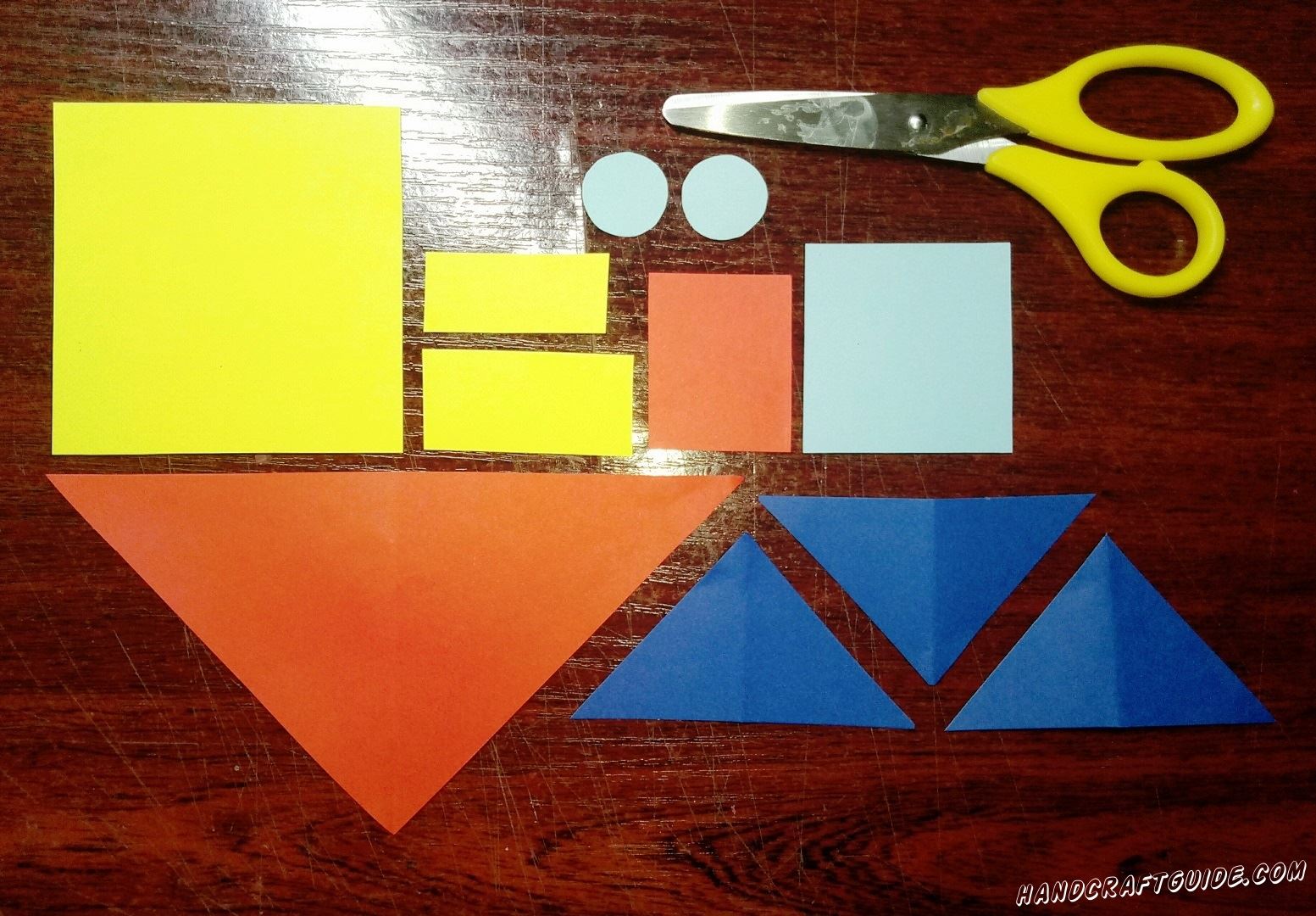 Для начала берем в руки бумаги, ножницы и вырезаем все необходимые нам фигурки: квадрат и два прямоугольника желтого цвета, три треугольника синего цвета, красный большой треугольник и небольшой квадрат, квадрат и два кружочка голубого цвета.