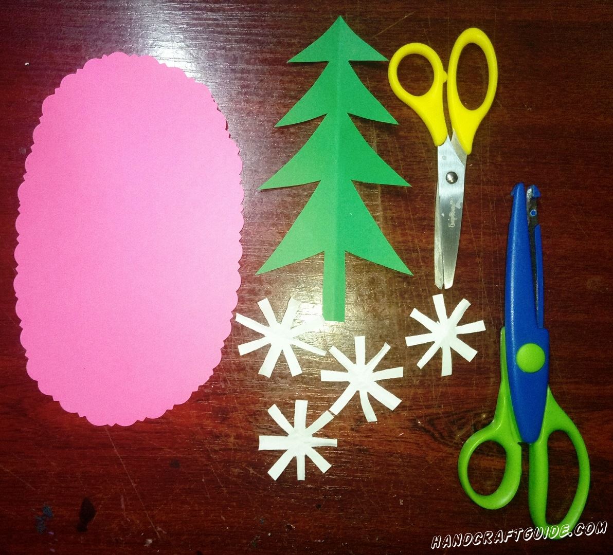 Вырезаем большой розовый овал из бумаги, и красиво делаем узоры по ободку. Также вырезаем зелёную ёлочку и четыре белых снежинки из бумаги.